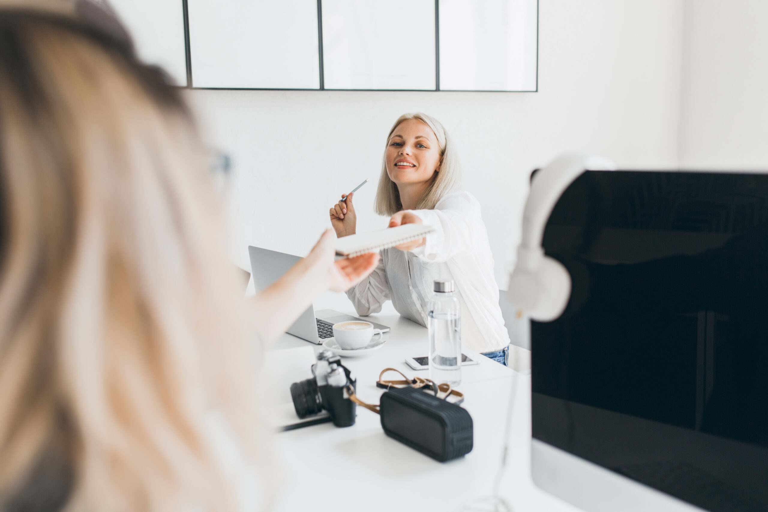 Smiling blonde secretary handed documents to colleague and drinking coffee. Fair-haired female programmer posing near computer with black screen while her friend enjoying latte.